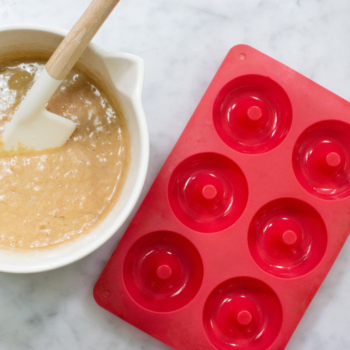 On a marble surface, the FarmSteady Doughnut Baking Pan, a red silicone mold with six cavities, awaits delicious homemade creations. Nearby, a white mixing bowl holds beige batter and has a spatula resting inside.