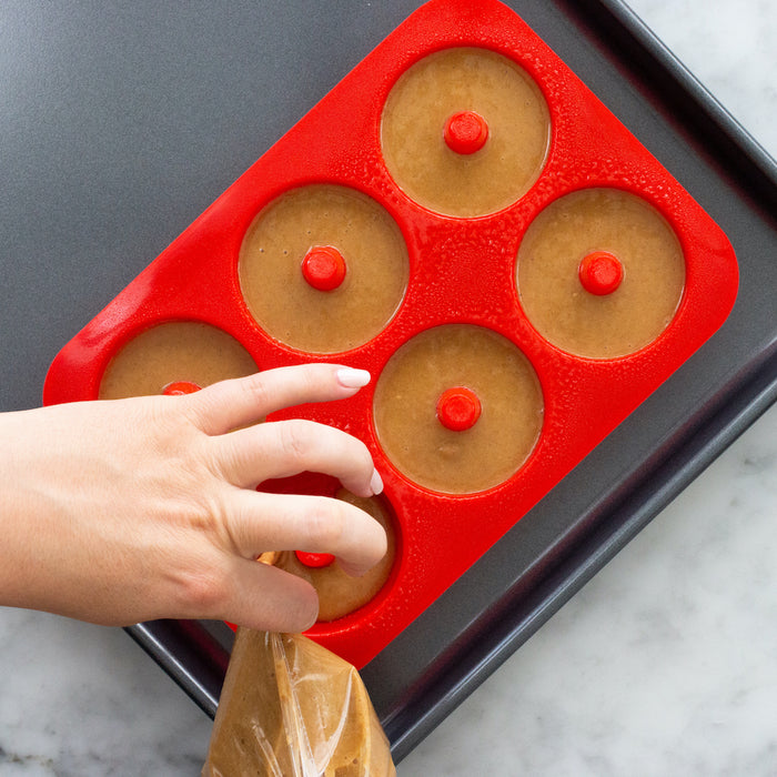 A hand fills a red FarmSteady Doughnut Baking Pan with rich brown batter using a piping bag. The pan sits on a sleek black tray on a pristine white marble countertop, ideal for making delicious homemade doughnuts.