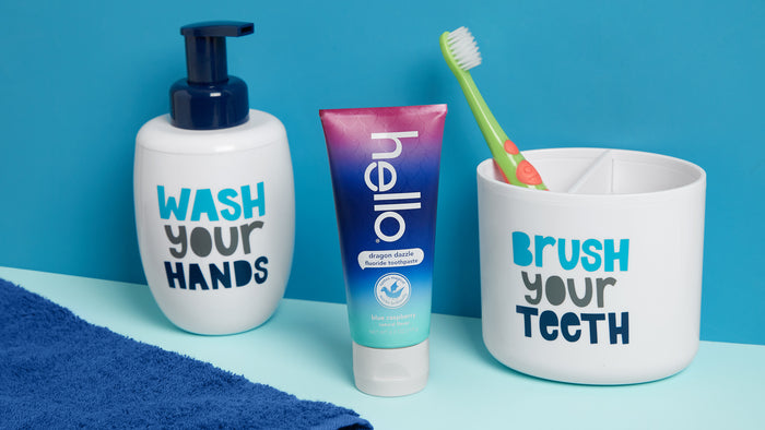 Bathroom setup with a Wash Your Hands soap dispenser, a Brush Your Teeth holder containing green and orange toothbrushes, hellos Dragon Dazzle Toothpaste with natural flavor, and a blue towel.