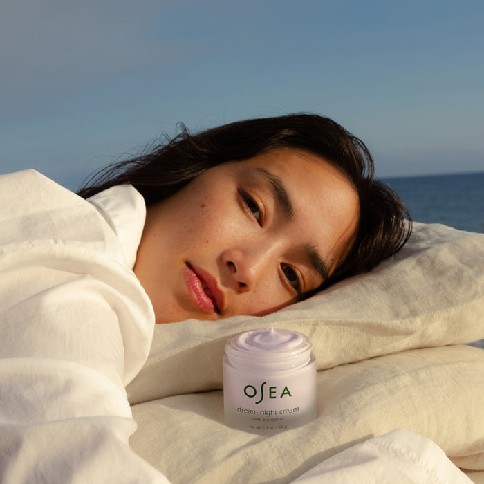 A woman in a white shirt rests her head on a pillow outdoors by the ocean, with OSEA Dream Night Cream with Bio-Retinol—an overnight moisturizer—placed in front of her under a clear blue sky.