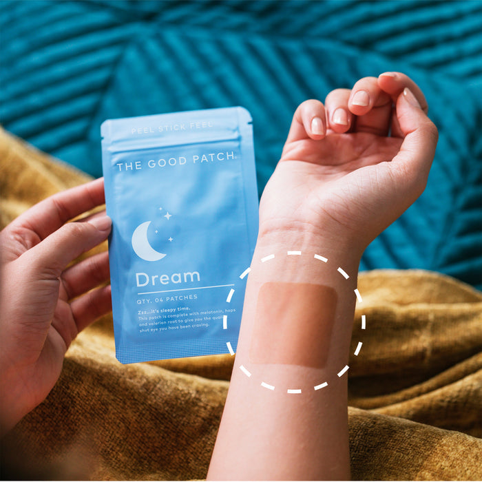 A person holds a blue pouch labeled The Good Patch: Dream Patch while wearing a sleep patch on their wrist. The quilted blue background emphasizes this innovative wellness product designed to support restful sleep.