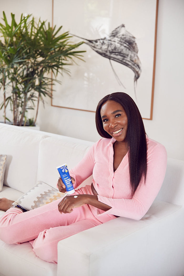In pink loungewear, a person smiles on a white couch, holding a blue tube of Asutras Dream the Night Away Melatonin Lotion, while a lush plant and abstract artwork enhance the backdrop.