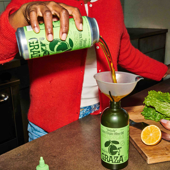 Wearing a red sweater, the person uses a funnel to pour Grazas Drizzle Extra Virgin Olive Oil - Refill from its recyclable green can into a bottle. A halved lemon and fresh lettuce are arranged beside the green oil bottle on the countertop.