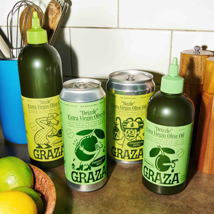 Four containers of Graza Drizzle Extra Virgin Olive Oil are on a counter, two in green squeeze bottles and two in metallic cans, featuring bird and orange illustrations. Kitchen utensils and limes enhance this elegant scene of recyclable refills.
