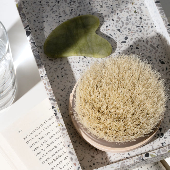 An EcoTools Dry Body Brush, round and beige, crafted from sustainable materials, and a green jade gua sha tool sit on a terrazzo tray beside a partially visible glass of water. Sunlight casts shadows on an open book, creating a minimalist and serene atmosphere.