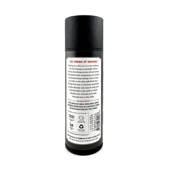 A black cylindrical Spinster Sisters Co. dry shampoo bottle features a white back label detailing its formula with deodorizing baking soda and volumizing rice starch, plus a barcode. The brand name and website are prominently displayed.