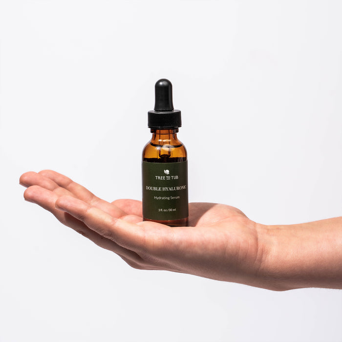 A hand holds a small amber glass bottle with a dropper labeled Dual Action Deep Hydrating Hyaluronic Acid Serum for Face by Tree To Tub against a plain white background. Perfect for hydrated skin, this serum refreshes and rejuvenates your complexion.