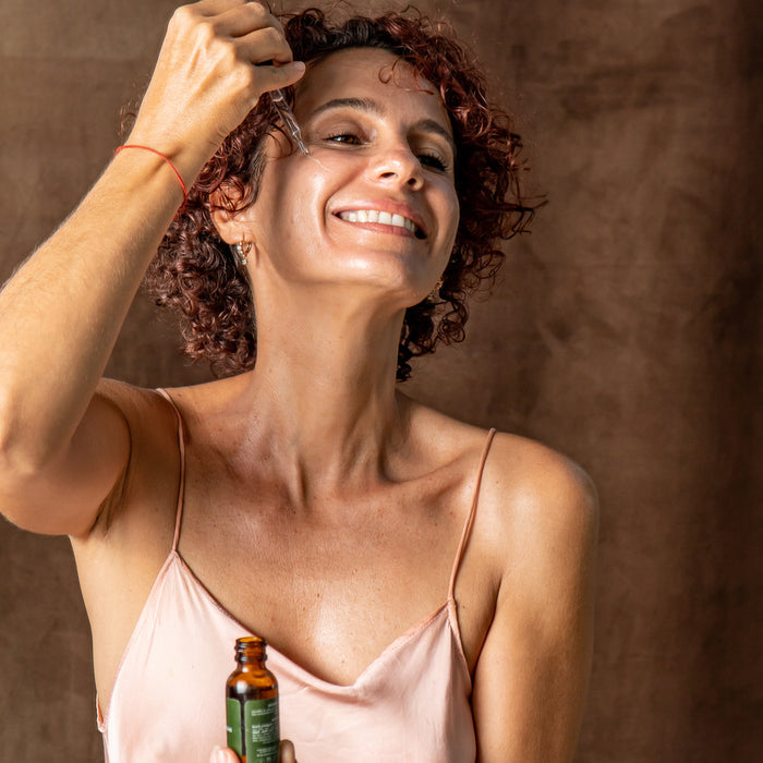 A woman with short curly hair smiles as she applies Tree To Tubs Dual Action Deep Hydrating Hyaluronic Acid Serum for Face with a dropper. She wears a light pink spaghetti strap top, her skin glowing with hydration against the soft brown background.