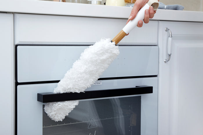 In a bright, clean kitchen with white cabinetry, someone uses a Full Circle Dust Whisperer Filament Microfiber Duster to clean the handle of the built-in oven door.