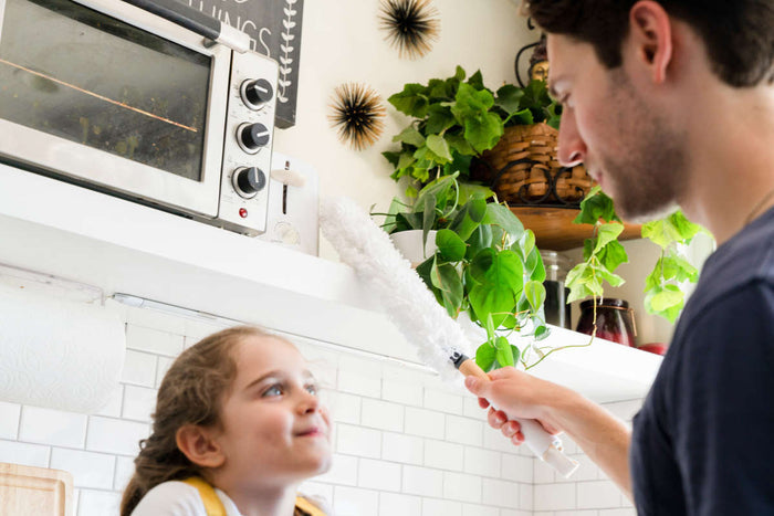 In the kitchen, a man playfully dusts a young girls hair with the Dust Whisperer Filament Microfiber Duster by Full Circle. The girl smiles, surrounded by plants and a compact oven on a shelf in the background.