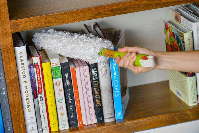 A hand delicately dusts a bookshelf filled with cookbooks using the Full Circle Dust Whisperer Filament Microfiber Duster. This green and yellow washable tool efficiently removes dust between the neatly arranged books on the wooden shelf unit.