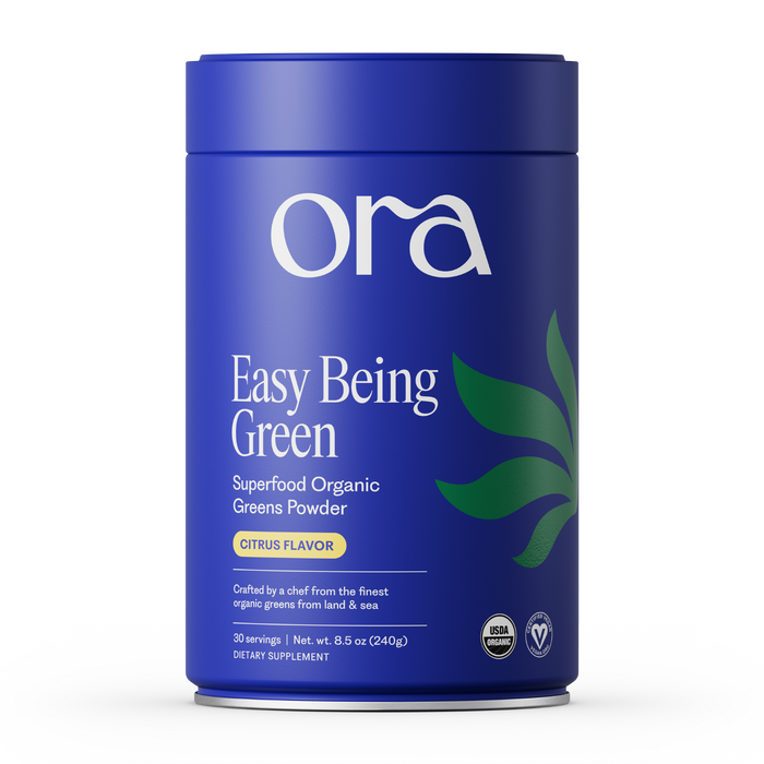 A blue cylindrical Ora Organic container labeled "Easy Being Green: Daily Active Superfood Greens Powder, Citrus Flavor," features green leaf graphics, certification icons, and highlights organic prebiotics on the front.