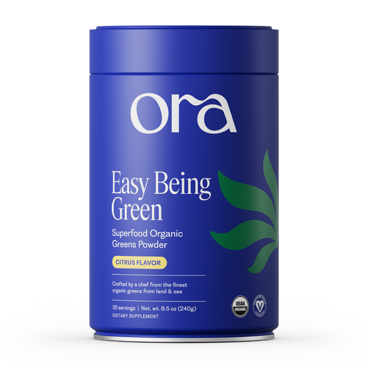 A blue cylindrical Ora Organic container labeled "Easy Being Green: Daily Active Superfood Greens Powder, Citrus Flavor," features green leaf graphics, certification icons, and highlights organic prebiotics on the front.
