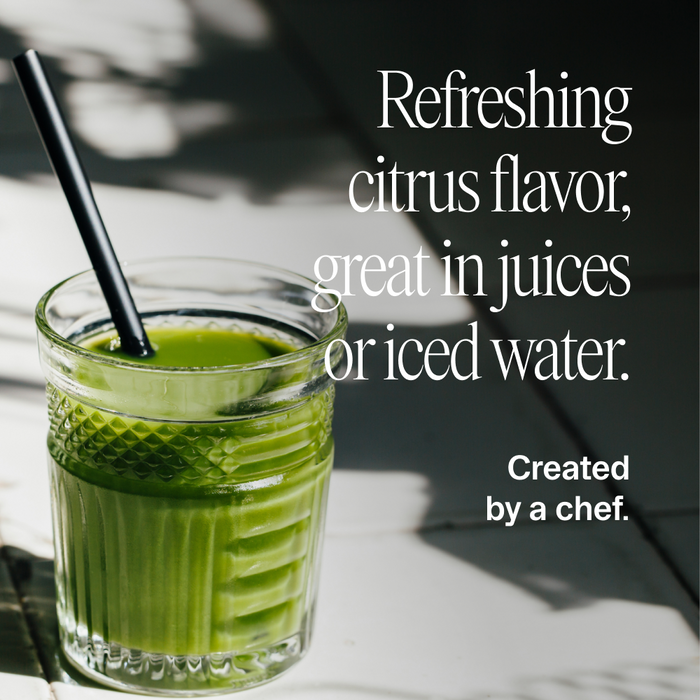 A glass of green juice with a black straw sits on a sunlit white surface. Text beside it reads: Easy Being Green by Ora Organic—delicious citrus taste, boosted with superfood greens. Perfect for juices or iced water. Chef-crafted.
