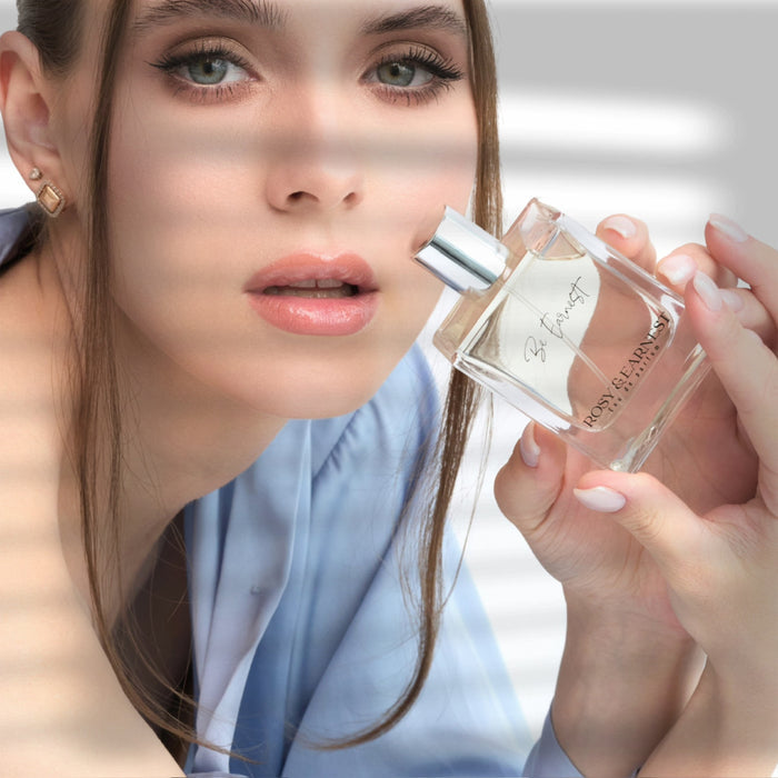 A woman with long hair, wearing a light blue shirt and natural makeup, holds a bottle of Rosy & Earnests Be Earnest perfume while looking at the camera. Sunlight casts striped shadows on her face and the cruelty-free, woody scent bottle she holds.