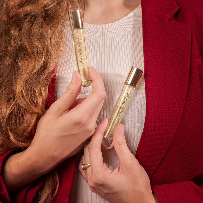 In a red blazer, a person with long hair holds two Pistachio Delight perfume vials by Rosy & Earnest, each with golden caps and elegant script. Wearing a cream ribbed top and a gold ring on their right hand, they exude the luxurious gourmand fragrance of Pistachio Delight.