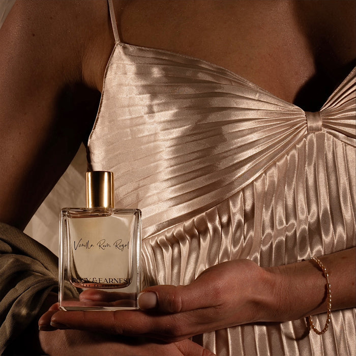 A person holds a Vanilla Rum Royal perfume bottle by Rosy & Earnest, featuring a gold cap and gourmand scent. Theyre in a shiny, champagne pleated dress, with a delicate gold bracelet. The warm, soft lighting highlights the cruelty-free allure of elegance.