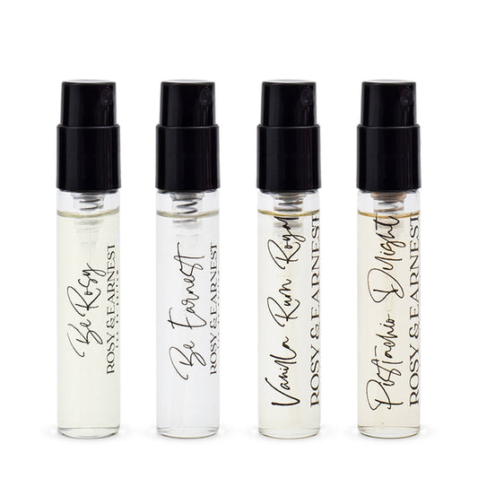 Four small spray bottles from the Rosy & Earnest Discovery Set are displayed in a row. Each has a black cap and labels with scent names: Be Rosy, Be Earnest, Vanilla Rum Rosy—featuring smooth vanilla and bold rum—and Perfumers Delight.