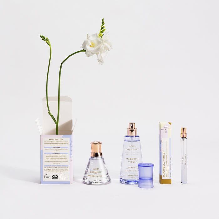 A display of Good Chemistrys Eau De Parfum Travel Spray, highlighting the vegan and cruelty-free Magnolia Violet scent. Various bottles and boxes are arranged around a tall white flower with a long stem in an open box, all set against a minimalist white background.