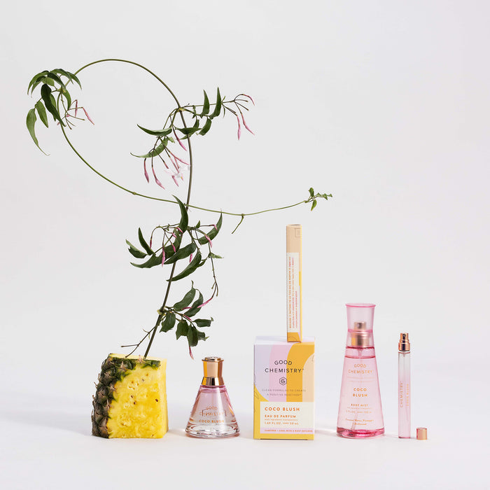 Four vegan perfume bottles and a box of Good Chemistry Eau De Parfum Travel Spray in Coco Blush scent are artfully arranged with half a pineapple and a leafy branch on a white background.