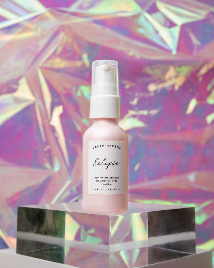 An Earth Harbor ECLIPSE Sheer Mineral Sunscreen SPF 30 bottle sits on a reflective glass cube, set against a background of colorful, iridescent patterns in pink, purple, and gold.
