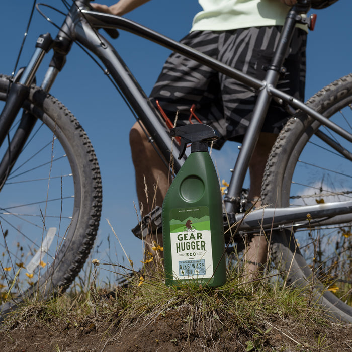 A person stands with a bicycle on a grassy hill under a clear blue sky. In the foreground, a green bottle labeled Gear Hugger Eco Bike Wash rests among wildflowers, emphasizing its gentle touch on nature and minimal environmental impact.