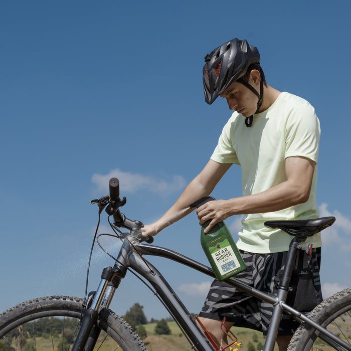 In an outdoor setting under a blue sky, a cyclist in a helmet and yellow shirt uses Gear Huggers Eco Bike Wash, a non-toxic green cleaner, to wash his black mountain bike, effectively minimizing environmental impact.