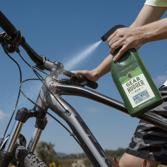 Under a clear blue sky, someone in camouflage shorts sprays Gear Huggers Eco Bike Wash onto a dusty bike. The bicycle with a dark frame and brand logo gleams as the eco-friendly cleaner minimizes environmental impact.