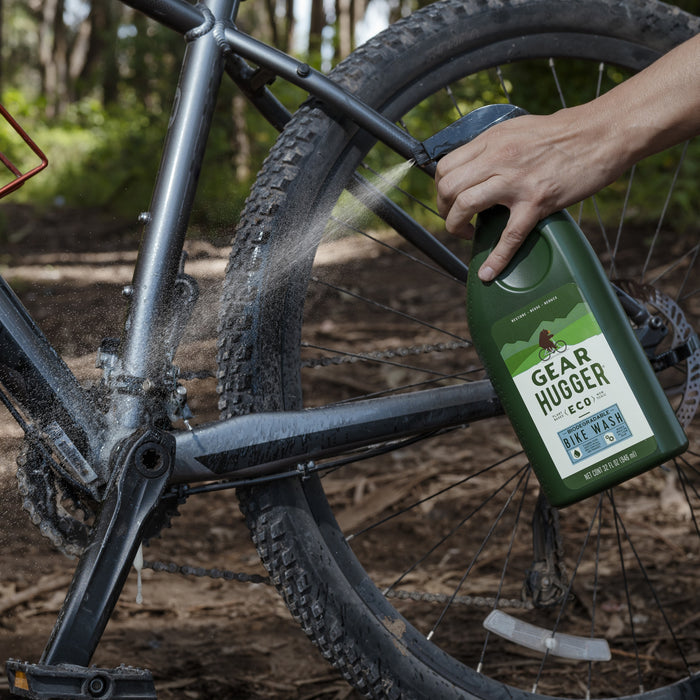 In a forest setting, someone non-toxically sprays their bike with Gear Hugger Eco Bike Wash. The green bottle, featuring a red gear icon, effectively cleans the frame while minimizing environmental impact and leaving the bike spotless.