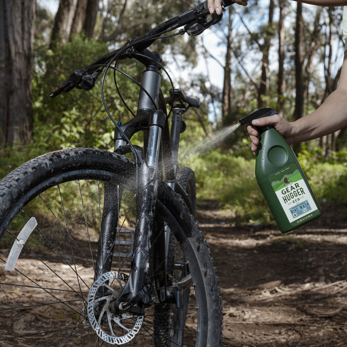 In a lush forest, someone uses Gear Huggers Eco Bike Wash on a muddy mountain bikes front wheel and suspension. Holding a green bottle labeled Non-toxic, they maintain their ride without worrying about environmental impact amid the surrounding trees and greenery.