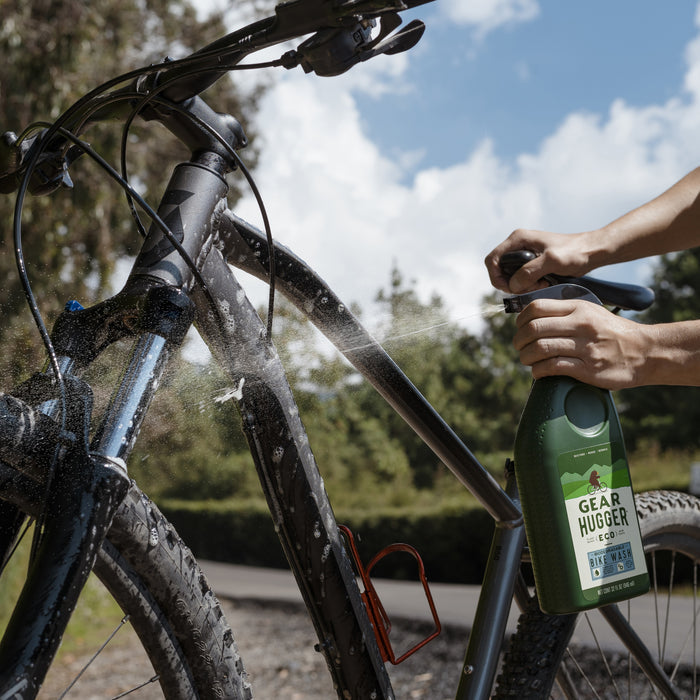 A person sprays Eco Bike Wash from a Gear Hugger bottle onto a black bike frame outdoors, with a cloudy sky and trees in the background on a sunny day.