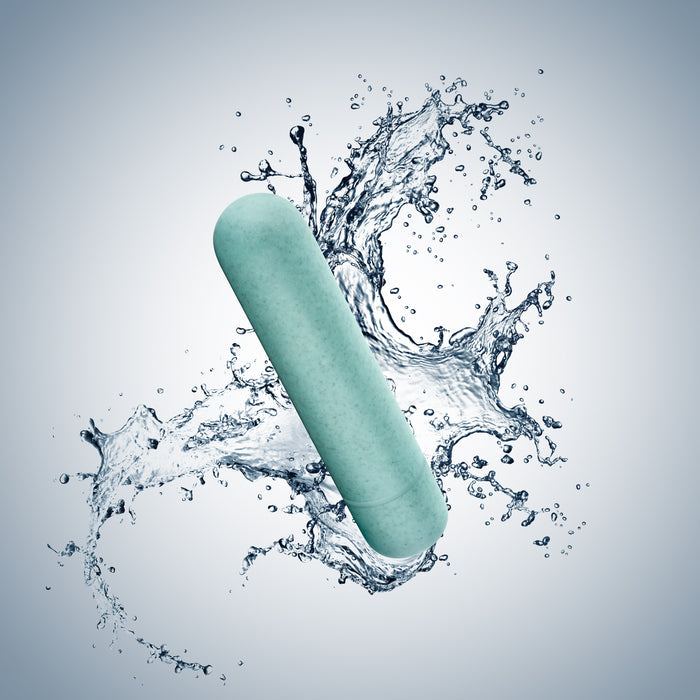 A teal capsule by Blush Gaias Eco Bullet hovers surrounded by splashes of water, with a light gray background. Dynamic curves emphasize this sleek, sustainable sex toys design.