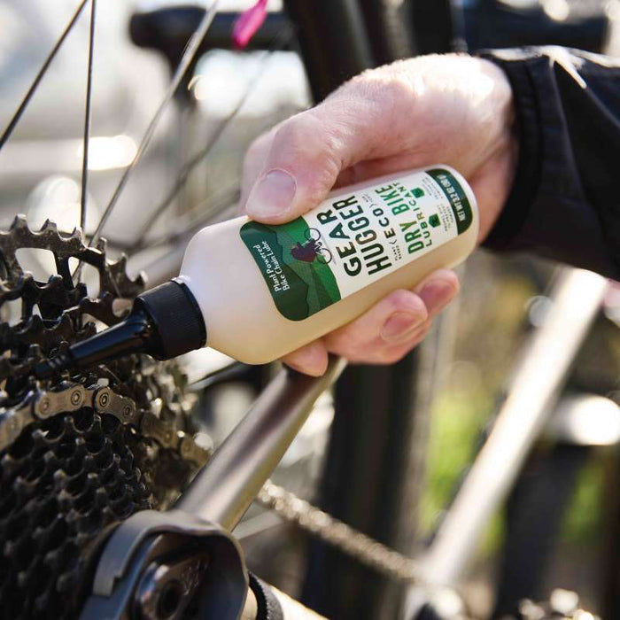 A person applies Gear Huggers Eco Dry Bike Lubricant to a bicycle chain near the rear derailleur. The bottles label is visible, with sunlight highlighting the metallic parts, emphasizing an eco-friendly touch on the chain and gears.