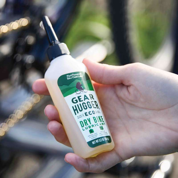 A hand holds a Gear Hugger Eco Dry Bike Lubricant with nozzle, showcasing its non-toxic, plant-powered formula. In the blurred background, a bike chain awaits this bio-based lubricant to work its eco-friendly magic.
