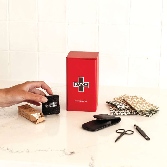A hand grabs a black patch from the Eco First-Aid Kit by PATCH, which contains a red box labeled PATCH, scissors, fabric swatches, hypoallergenic bandages, a pen, and a small black case—all displayed on white marble with a tiled background.
