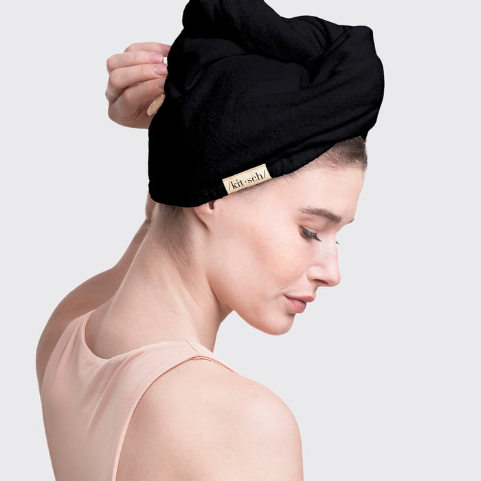 A light-skinned person, in a sleeveless peach top, is seen sideways. Their hair is wrapped in a Kitsch Eco-Friendly Hair Towel made from organic cotton with a black turban style. The background is white.