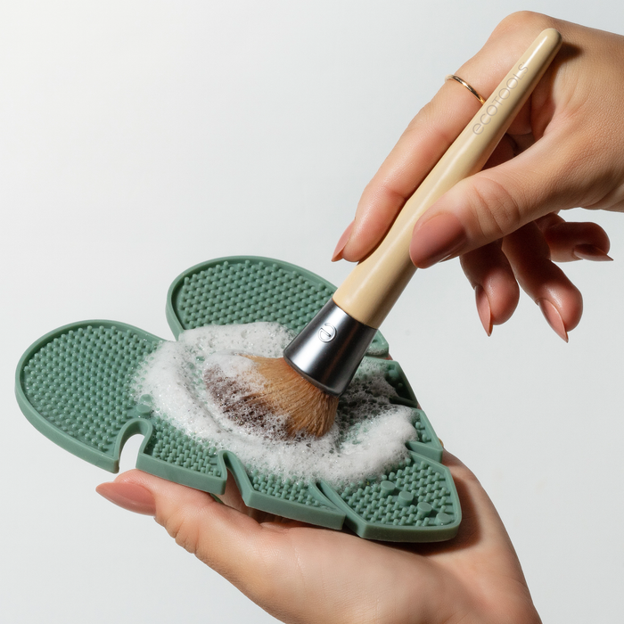 Hands diligently clean a makeup brush, featuring a light wooden handle and metal ferrule, on the EcoTools Eco Makeup Brush Cleansing Pad. The textured green silicone pad ensures effective cleansing with cruelty-free soap while keeping your tools pristine and environmentally friendly.