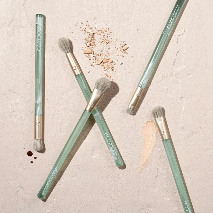 The EcoTools Eco Sea Gems Eye Set includes five makeup brushes with green handles. They are scattered on a textured beige surface, accompanied by makeup smudges and powder crumbs. Four cruelty-free, vegan brushes lie in various orientations, while one is positioned horizontally at the top.