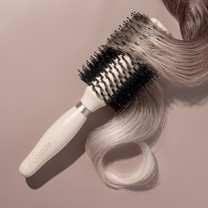 The EcoTools Eco Volumizing Round Brush, with a wooden handle and vegan boar bristles, holds long wavy light brown hair beautifully against a soft beige backdrop, highlighting its bamboo craftsmanship.