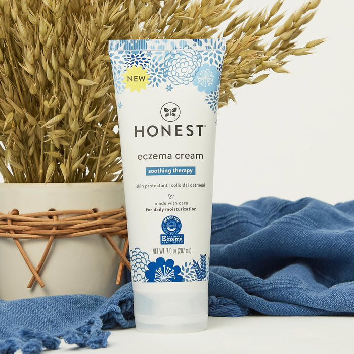 The Honest Companys Eczema Relief Cream is pictured with wheat stalks, featuring soothing colloidal oatmeal. A nearby blue cloth highlights its suitability for daily moisture on eczema-prone skin.
