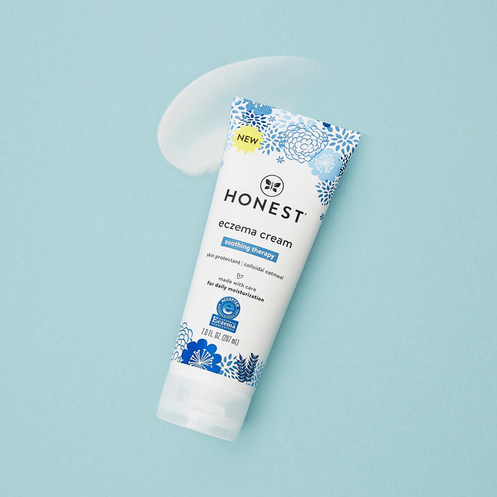 A tube of The Honest Companys Eczema Relief Cream sits on a light blue background, designed for eczema-prone skin. Next to it is a small smear, and the packaging features blue floral designs highlighting soothing therapy and daily moisture benefits.