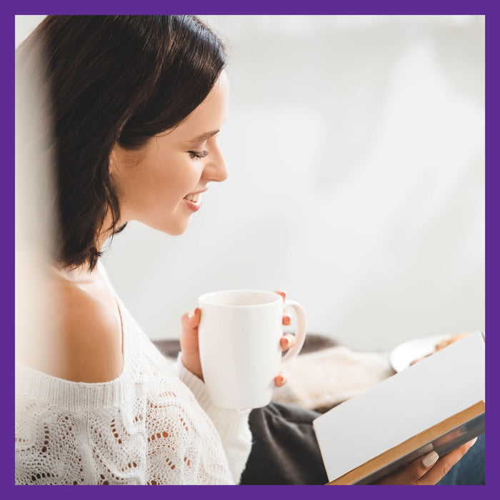 A woman with shoulder-length dark hair smiles while holding a white mug and reading a New Chapter book on immune support. She wears a white off-the-shoulder sweater, with the blurred background subtly hinting at Elderberry Gummies, Organic Whole-Food Gummies for Immune Support beside her.