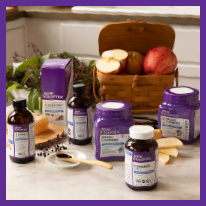 A variety of health supplement bottles and jars with purple labels are arranged on the countertop. New Chapter Elderberry Gummies, known for their antioxidant action, accompany a wooden basket of perfectly sliced apples beside a small dish with a spoon.