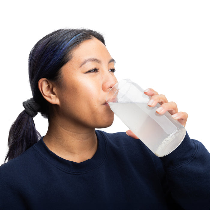 A person with a ponytail drinks from a clear glass filled with water enhanced by Drinklits Electrolyte Hydration Tablets. Wearing a navy blue sweater, they appear relaxed with slightly closed eyes against the plain white background.