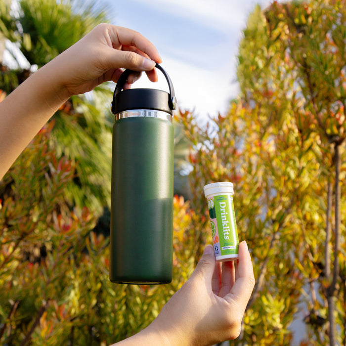 Two hands hold a green insulated bottle and a small cylindrical container labeled Drinklits, featuring a green label advertising zero sugar. The backdrop of lush greenery and clear blue sky suggests the refreshing electrolytes in the Electrolyte Hydration Tablets.