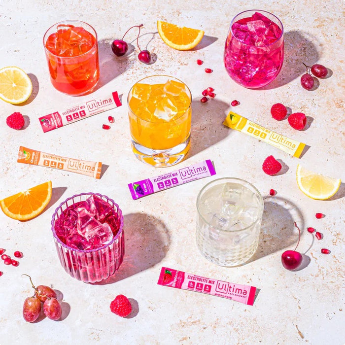 A vibrant array of colorful drinks in glasses with citrus, cherries, raspberries, and grapes surround Ultima Replenishers Electrolyte Mix Variety Pack - For Hydration packets on a light speckled surface, promising optimal hydration with every sugar-free sip.