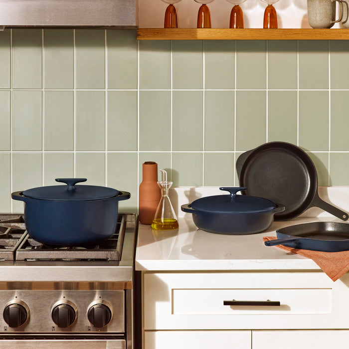 In a kitchen with light green tile backsplash, a stove features the Caraway Cast Iron Dutch Oven with Flat Lid, 6.5 qt in blue, noted for heat retention. The countertop has two non-toxic pots and olive oil, while a wooden shelf displays bottles and a plant.