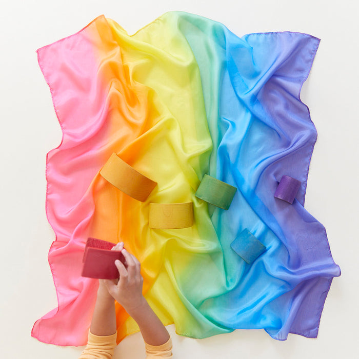 On a vibrant Enchanted Playsilk from Sarahs Silks, stretching in pink to purple hues, six matching cylindrical containers invite imaginative play. In the scenes lower left corner, hands cradle a red container.