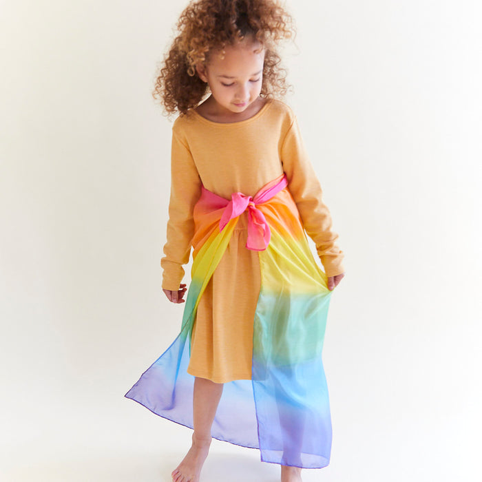 A child in a mustard dress with long curly hair gazes down, wearing Sarahs Silks Enchanted Playsilk tied at the waist. Made from mulberry silk, it flows elegantly to the side against a plain white background, ideal for inspiring open-ended play.