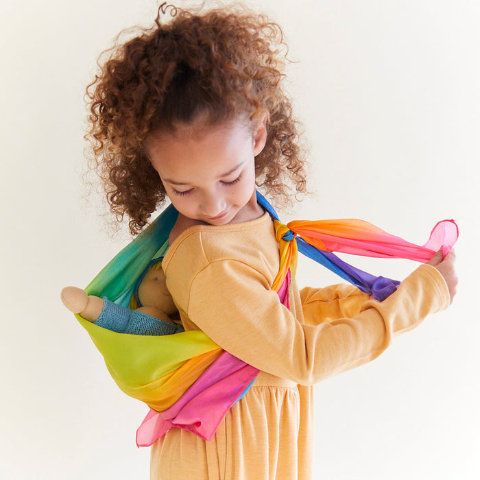 A young girl with curly hair in a yellow dress carries a doll in an Enchanted Playsilk sling by Sarahs Silks, tenderly looking down as she holds the ends. A plain light background highlights this imaginative, open-ended play moment.